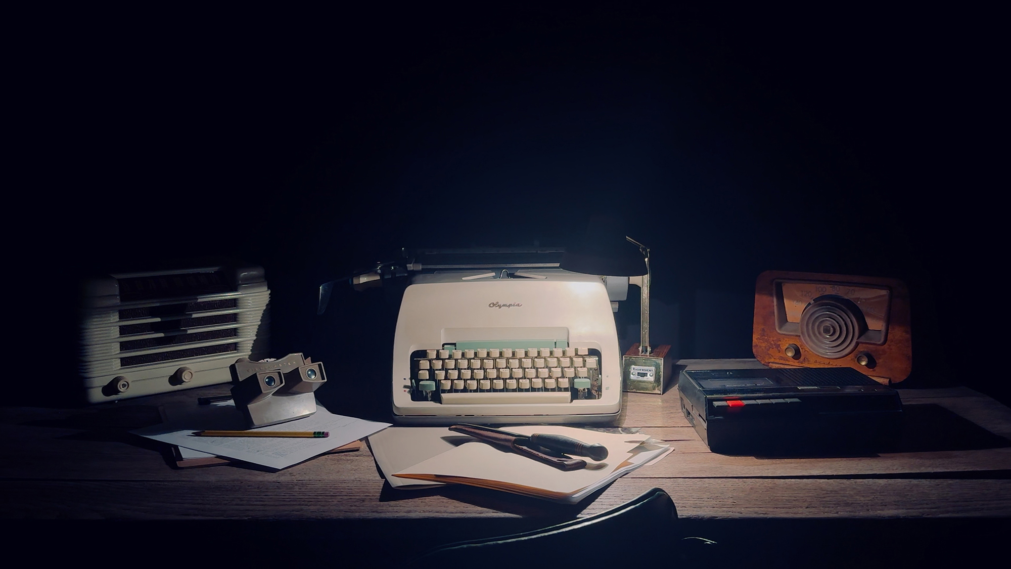 an old wooden desk with a typewriter, a viewfinder, a stack of papers, a file foolder, a knife, a tape deck, a radio, and a lamp